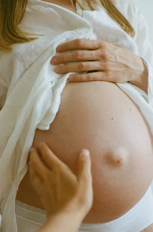 Ventre de femme enceinte en gros plan, caressé par une main et couvert d’une chemise blanche entrouverte.