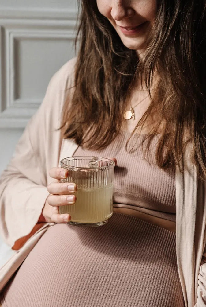 Femme enceinte souriante tenant une boisson au gingembre dans une tasse en verre – complément naturel contre les nausées de grossesse.