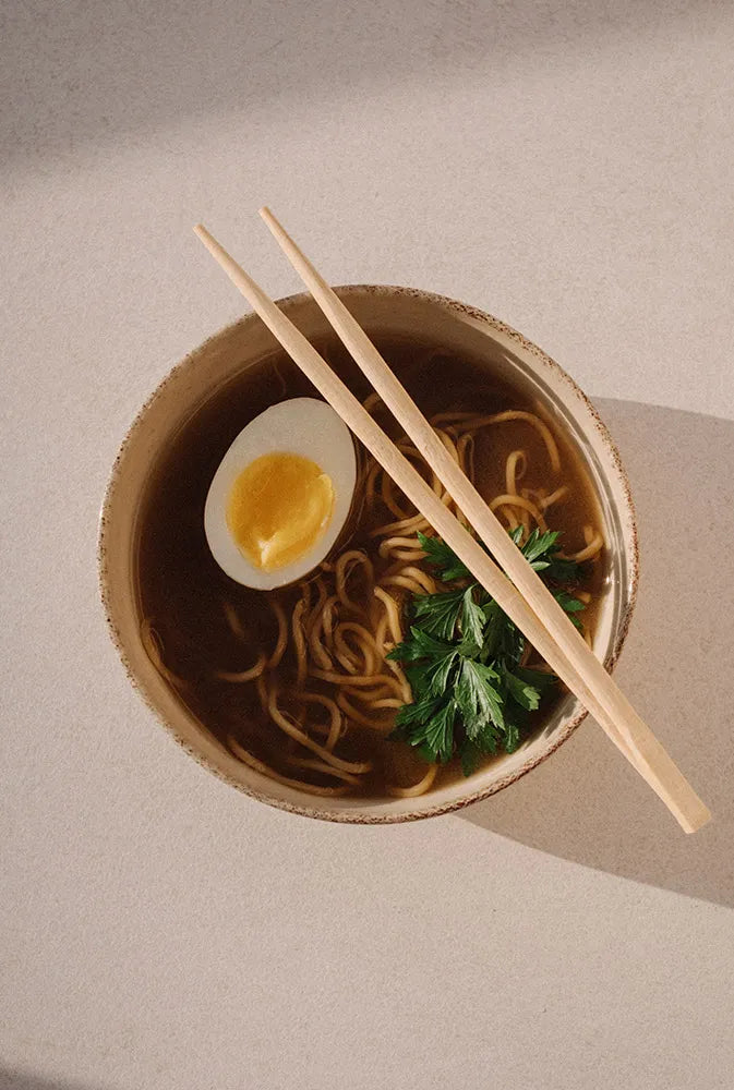 Bol de bouillon fumant aux nouilles, œuf dur et herbes fraîches, avec baguettes posées – préparation saine et réconfortante à base de Broth, idéale pour la récupération post-partum et l’hydratation.