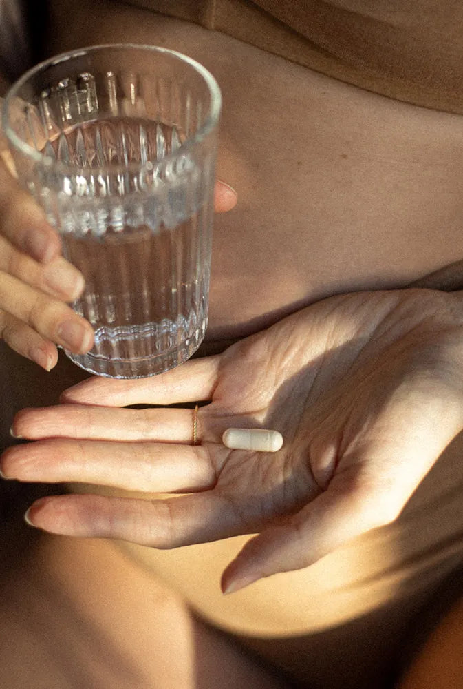 Main posée sur un ventre de femme tenant un verre d’eau et une gélule – complément alimentaire pensé pour les besoins spécifiques des femmes, entre équilibre hormonal, énergie et bien-être global.