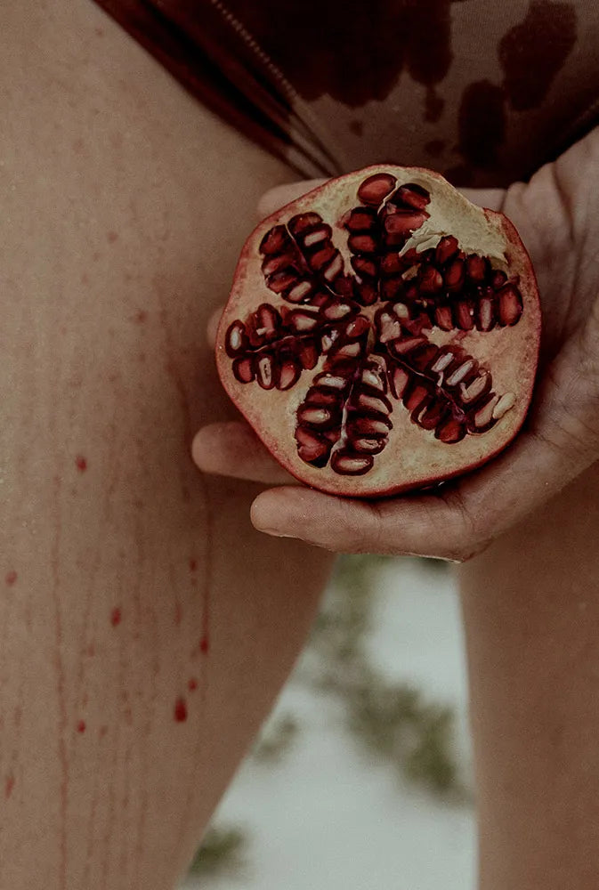 Main féminine tenant une grenade éclatée, avec des gouttes évoquant le cycle menstruel – image symbolique et puissante autour des règles, du féminin sacré et du soutien naturel pendant les menstruations.